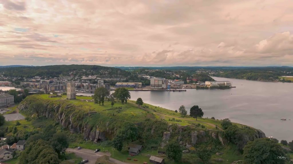 Aerial view of historic stone tower on grassy hill, overlooking water and cityscape with greenery.