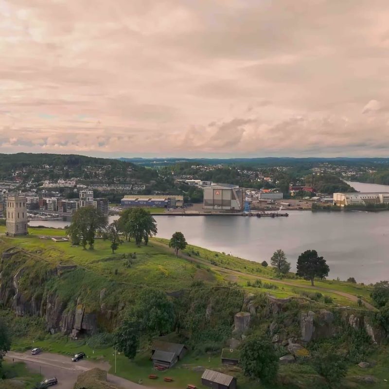 Aerial view of historic stone tower on grassy hill, overlooking water and cityscape with greenery.
